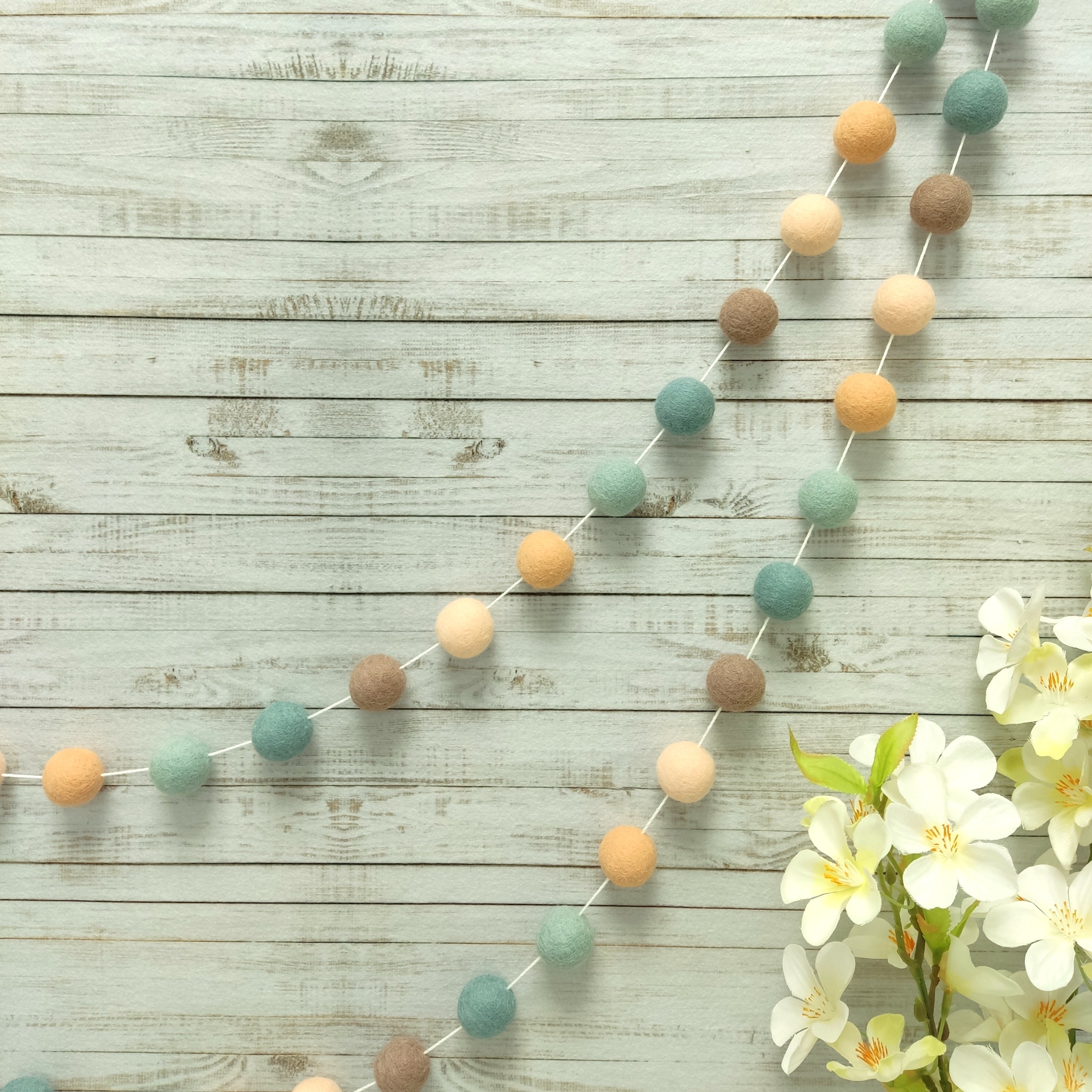 Guirlande de pompons feutrés sauge pêche suspendue sur un mur, teintes beige rosé, orange pêche, beige cappuccino, bleu nordique et vert céladon