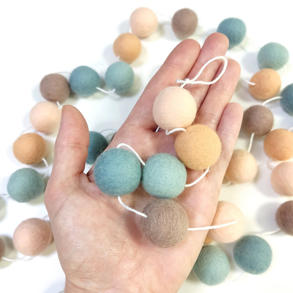 Zoom sur les pompons en laine mérinos sauge pêche dans une main, teintes naturelles et apaisantes