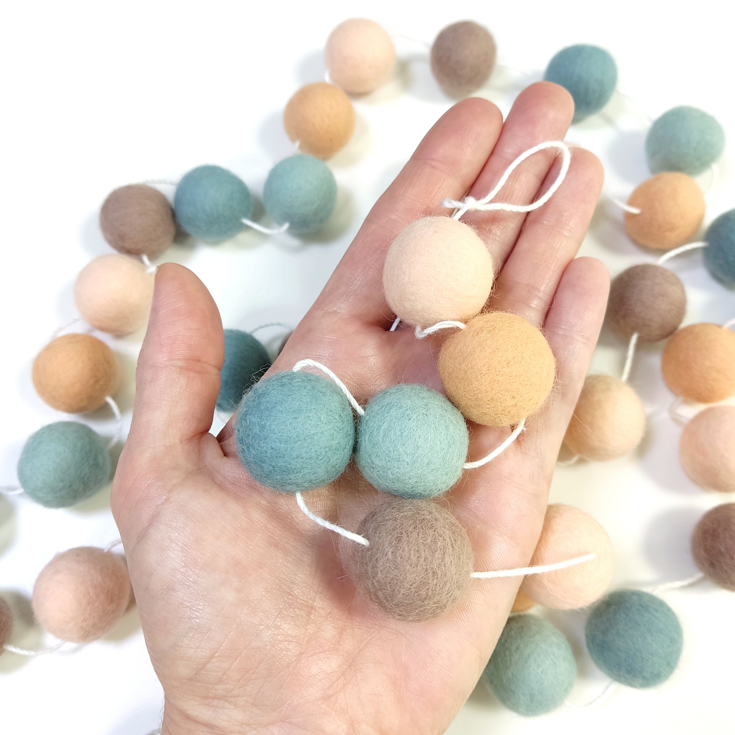 Zoom sur les pompons en laine mérinos sauge pêche dans une main, teintes naturelles et apaisantes