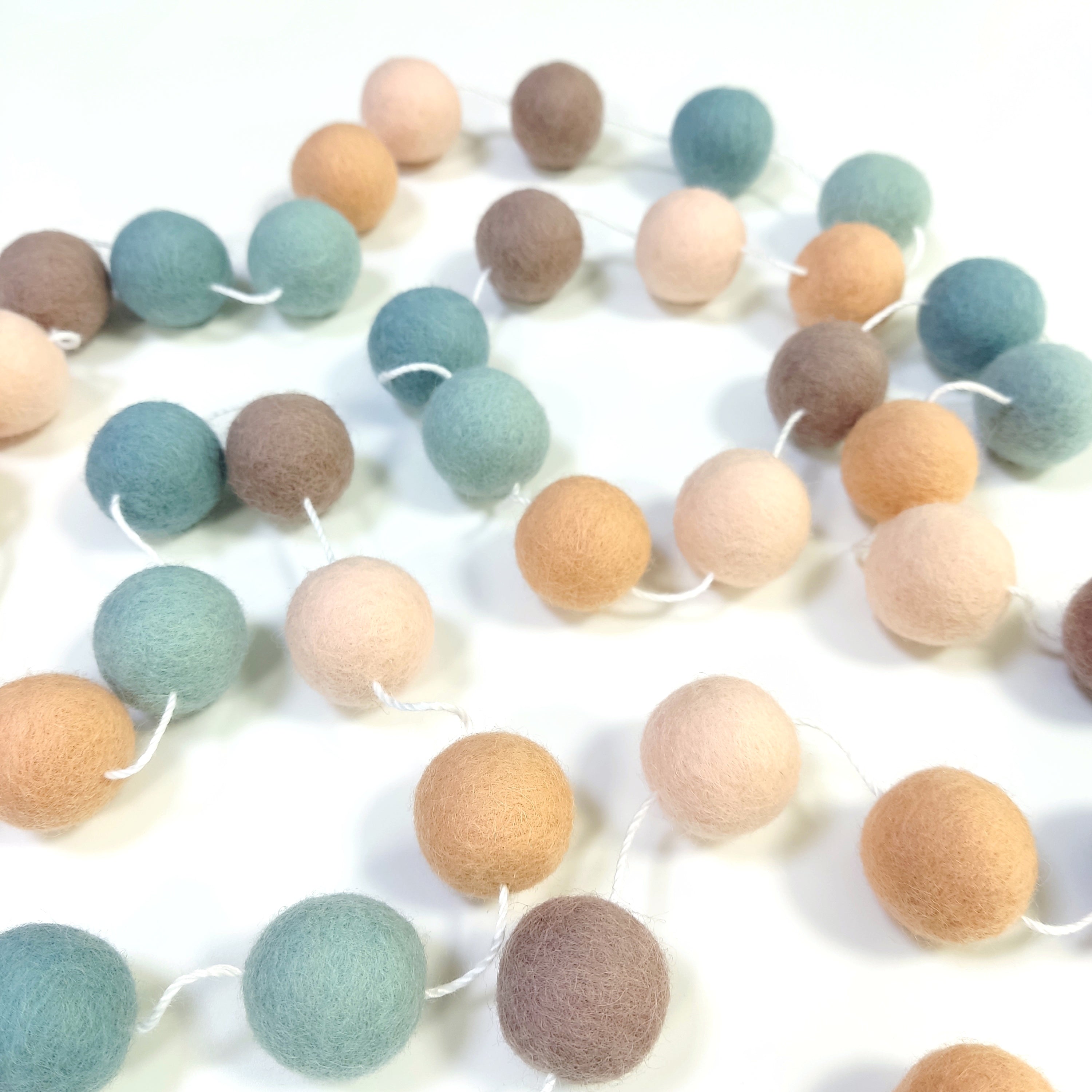 Guirlande de pompons feutrés sauge pêche étalée sur une table avec les pompons espacés sur le fil