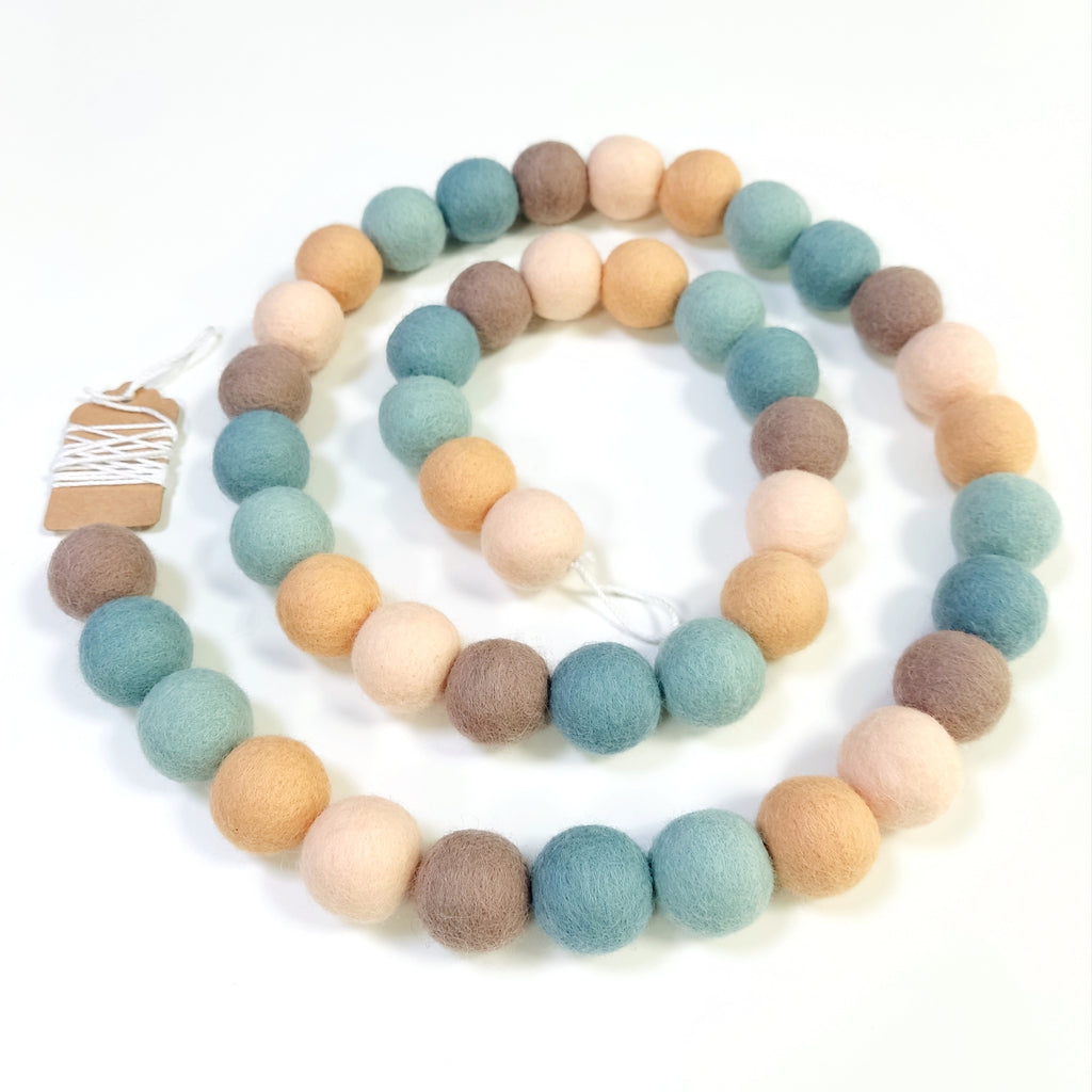 Guirlande de pompons feutrés sauge pêche disposée en spirale sur fond blanc
