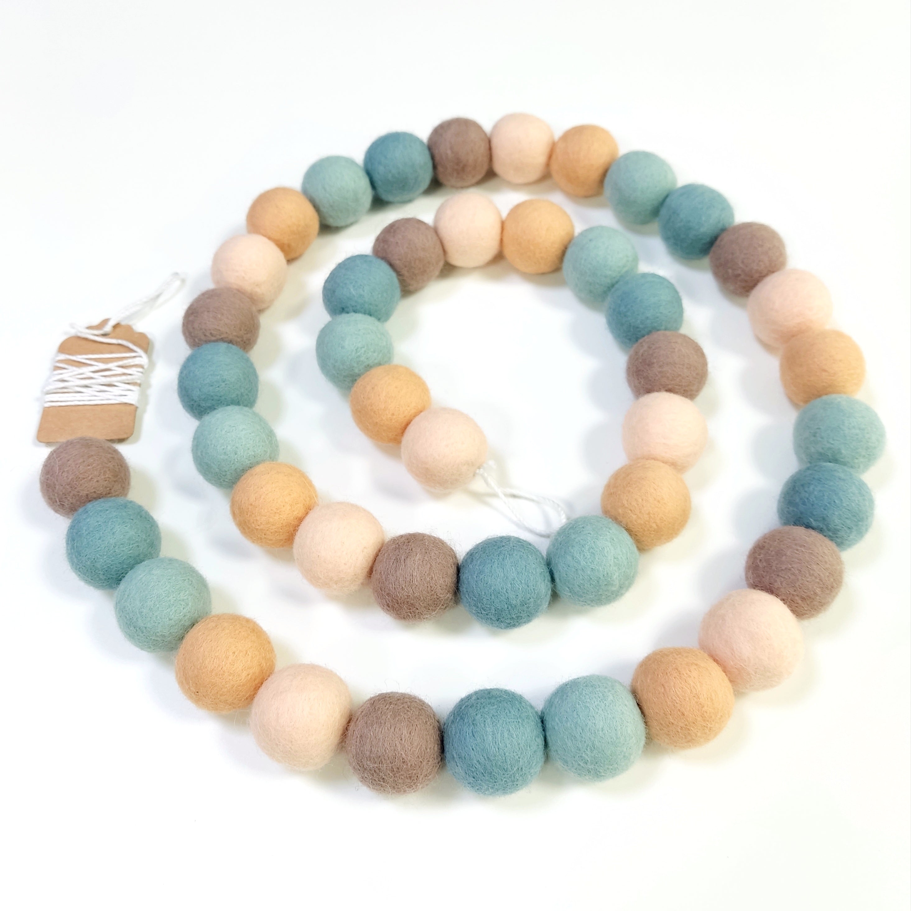 Guirlande de pompons feutrés sauge pêche disposée en spirale sur fond blanc