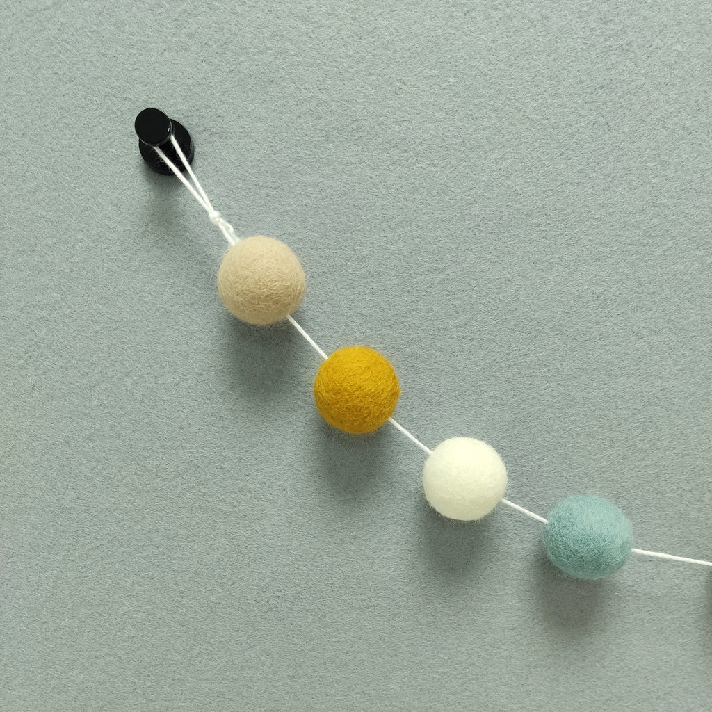 Zoom sur les 4 couleurs de la guirlande de pompons en feutre scandinave : bleu, ocre, blanc, beige poudré