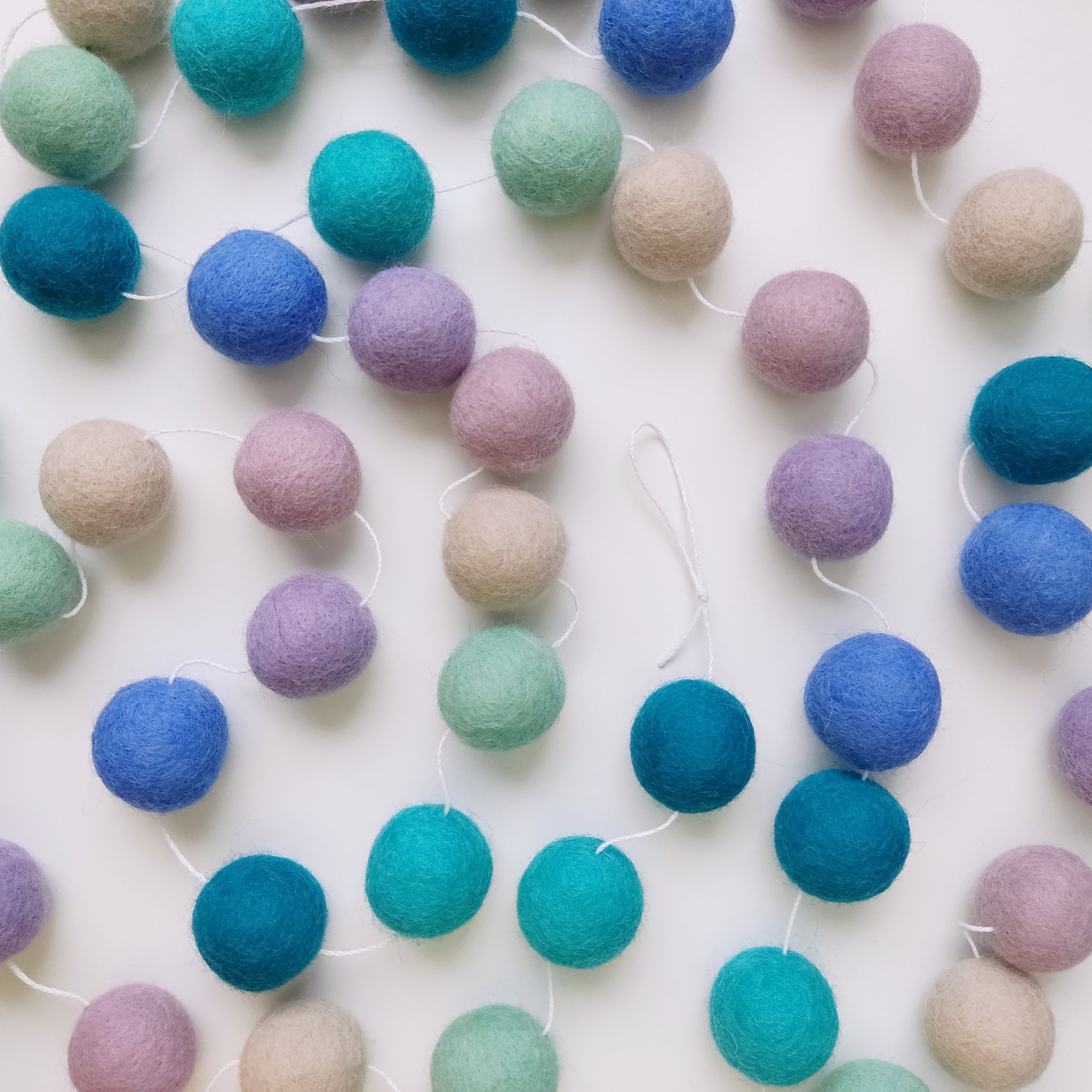 Guirlande de pompons en laine feutrée couleurs sirène étalée sur une table