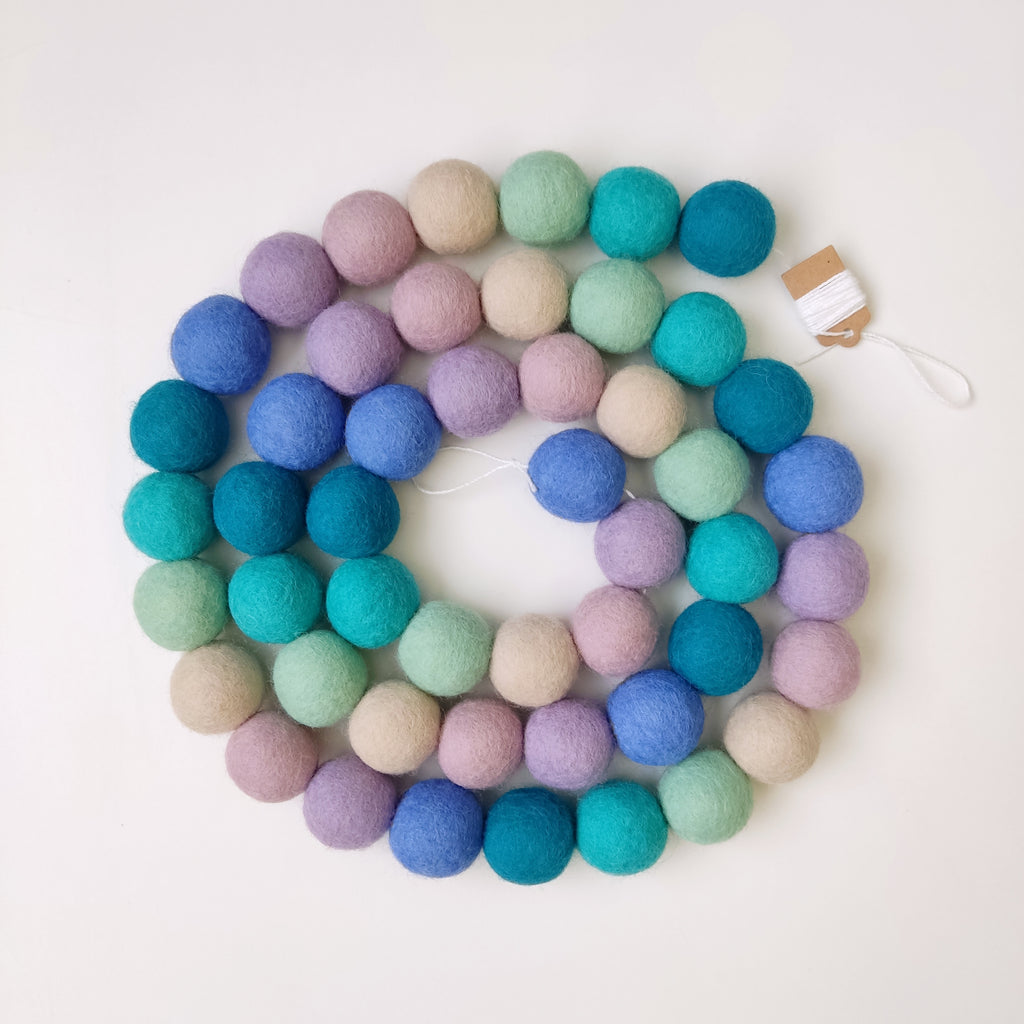 Guirlande de pompons sirène bleu mauve turquoise disposée en spirale sur une table vue de dessus