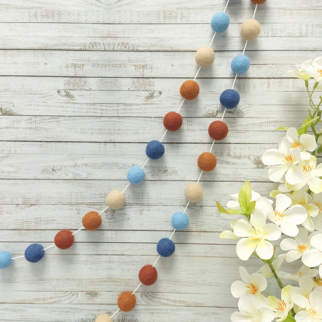 Guirlande de pompons feutrés aux teintes terracotta et bleu suspendue sur un mur