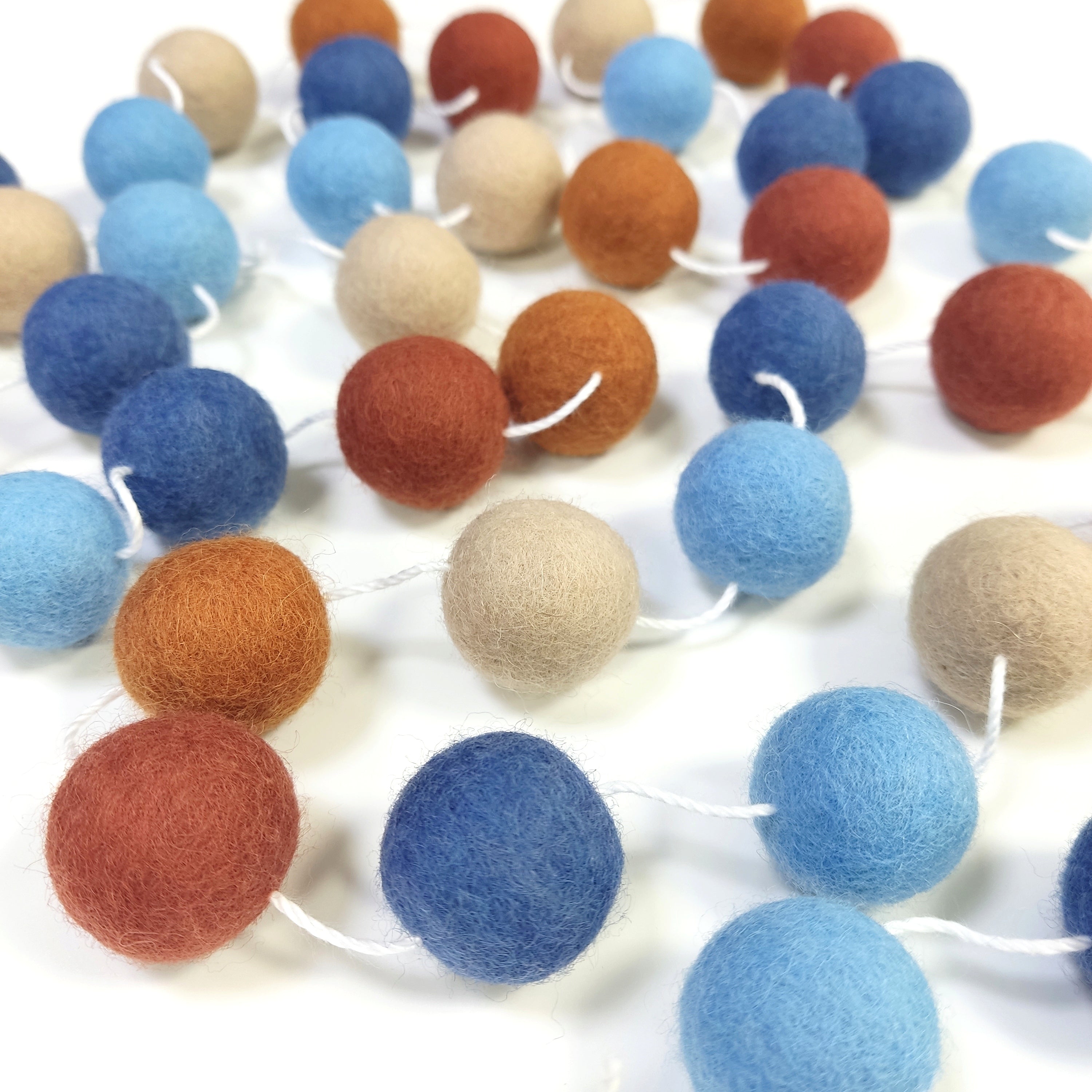 Guirlande de pompons en laine feutrée terracotta, beige et bleu étalée sur une table