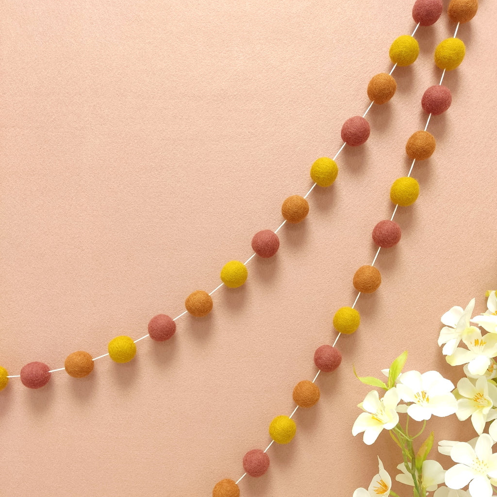 Guirlande de pompons feutrés terracotta ocre suspendue sur un mur, teintes terracotta rosé, terracotta cuivré et ocre doré