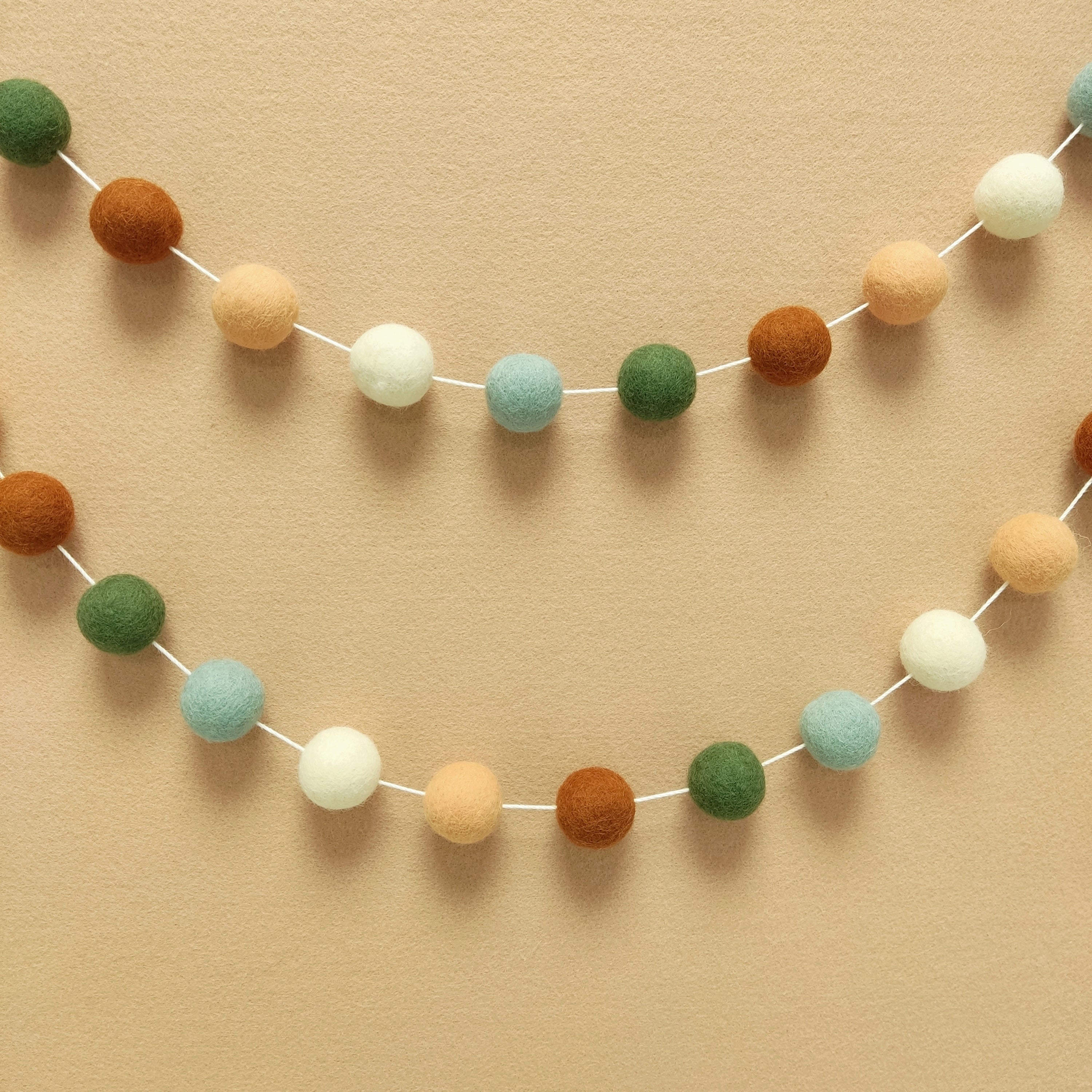 Guirlande de pompons feutrés terracotta, vert céladon et blanc suspendue en arc de cercle sur un mur, faite main en France