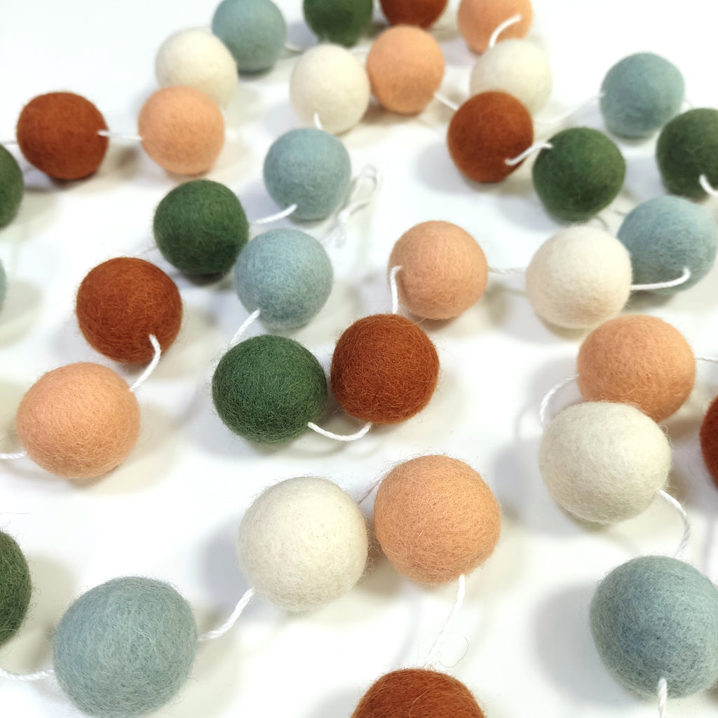 Guirlande en boules de feutre terracotta sauge blanc étalée sur une table avec les pompons espacés