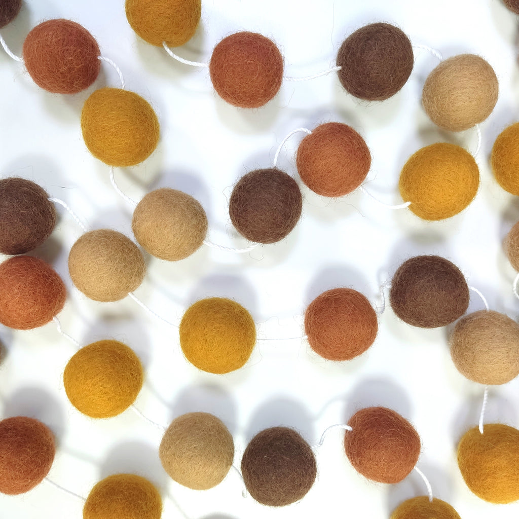 Guirlande de pompons en laine feutrée ocre, marron et terracotta étalée sur une table