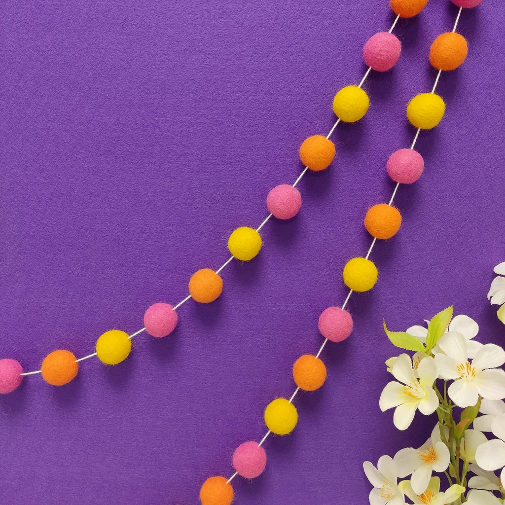 Guirlande de pompons feutrés tutti frutti suspendue sur un mur, teintes rose bonbon, orange carotte et jaune soleil