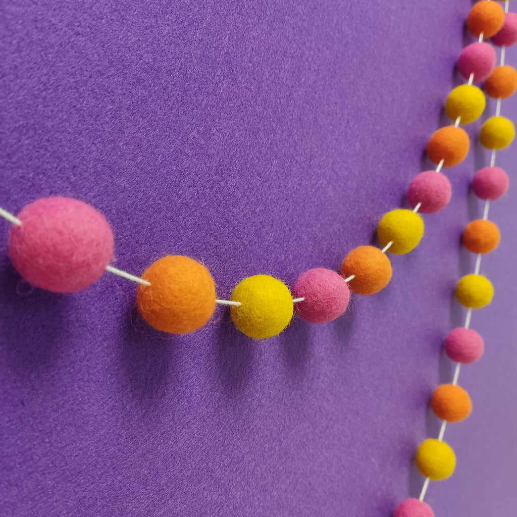 Guirlande de pompons feutrés aux teintes festives rose bonbon, orange carotte et jaune soleil vue de profil