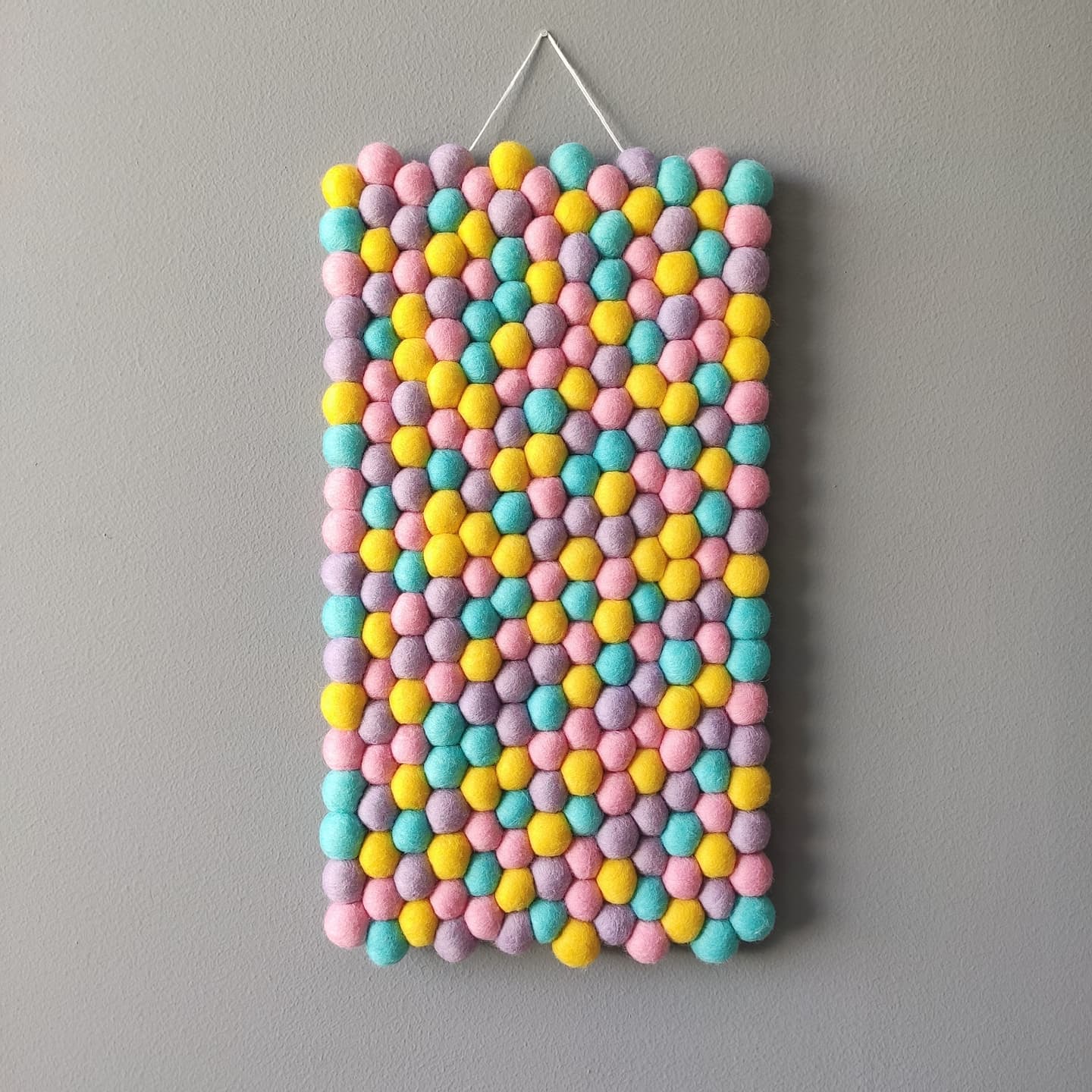 Tapisserie murale pixel pastel en pompons de feutre multicolores rose dragée, jaune citron, bleu lagon et mauve, suspendue par un cordon blanc