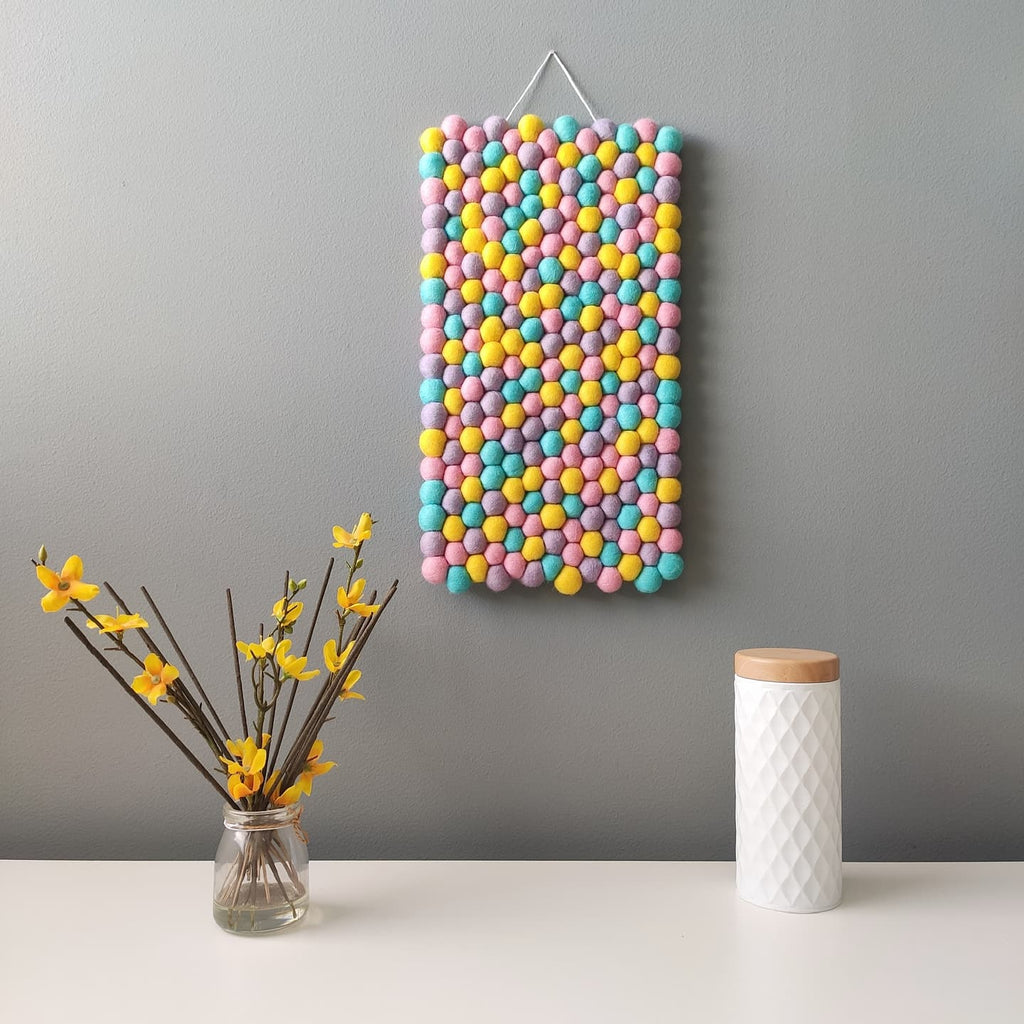 Tapisserie décorative en pompons de feutre pastel accrochée sur mur gris, avec vase de fleurs jaunes et pot blanc